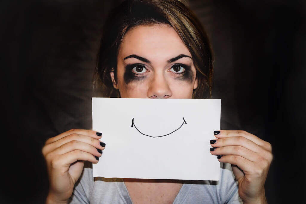 A girl with smudged mascara holding a paper with a smile drew on it over her face 