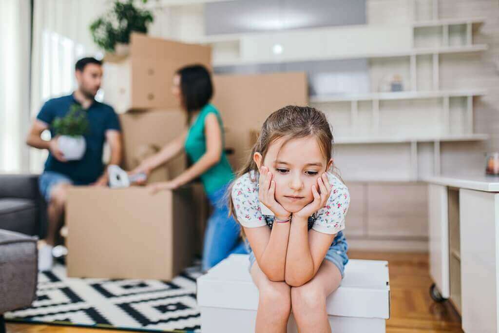 A sad girl leaning on the box while her mom and dad are packing behind her