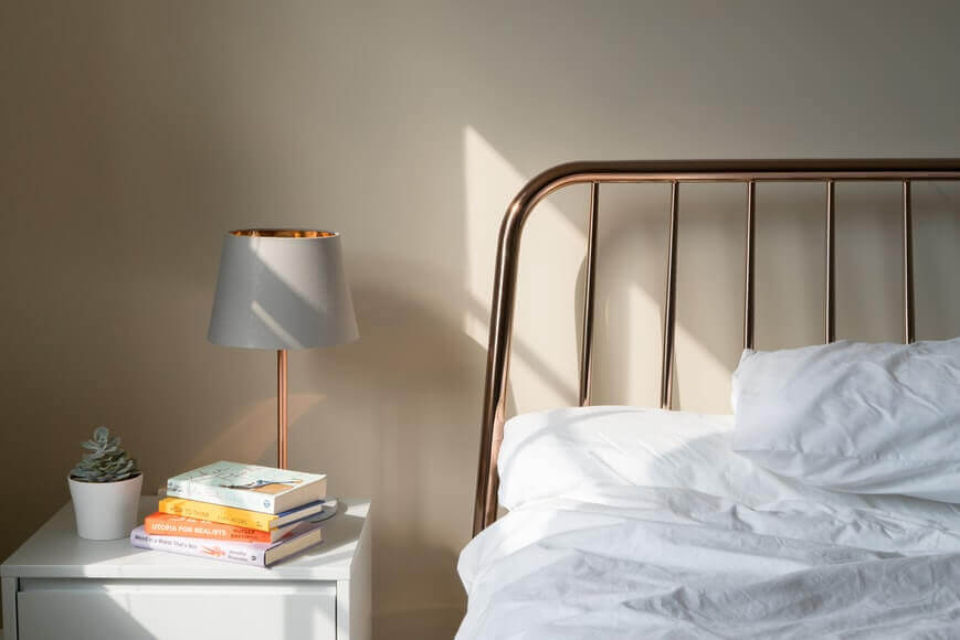 A corner of the bed, nightstand on the left, with lamp, books, and a plant