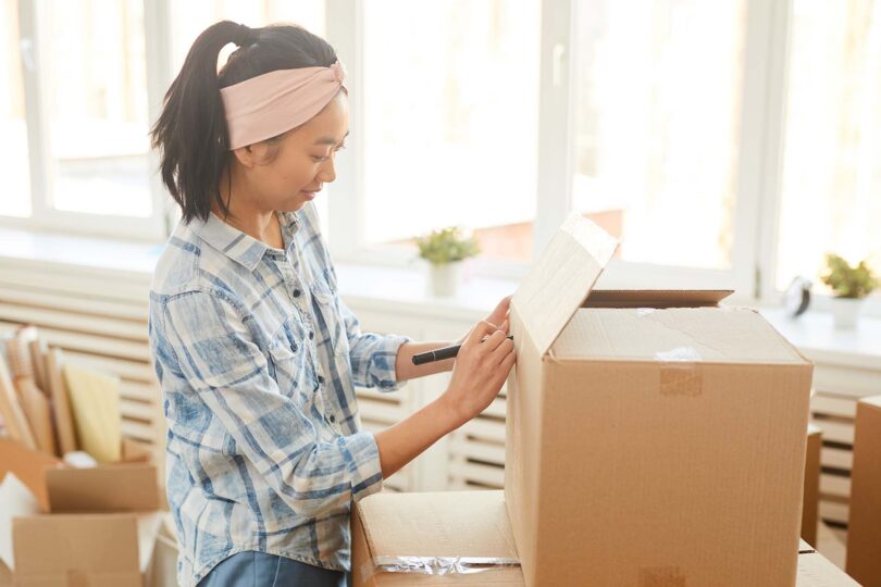 Side view portrait of young Asian woman signing cardboard boxes in sunlight while moving out to new house or apartment, copy space
