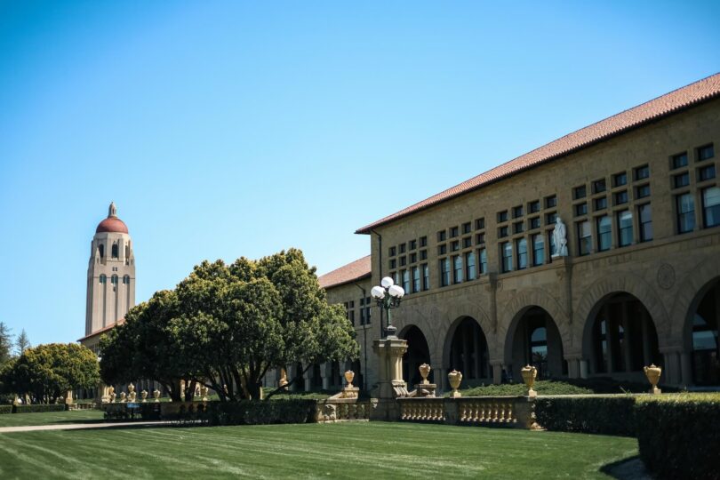 Historic sandstone buildings on the Stanford University campus featuring a tall clock tower in the background under a clear sky.